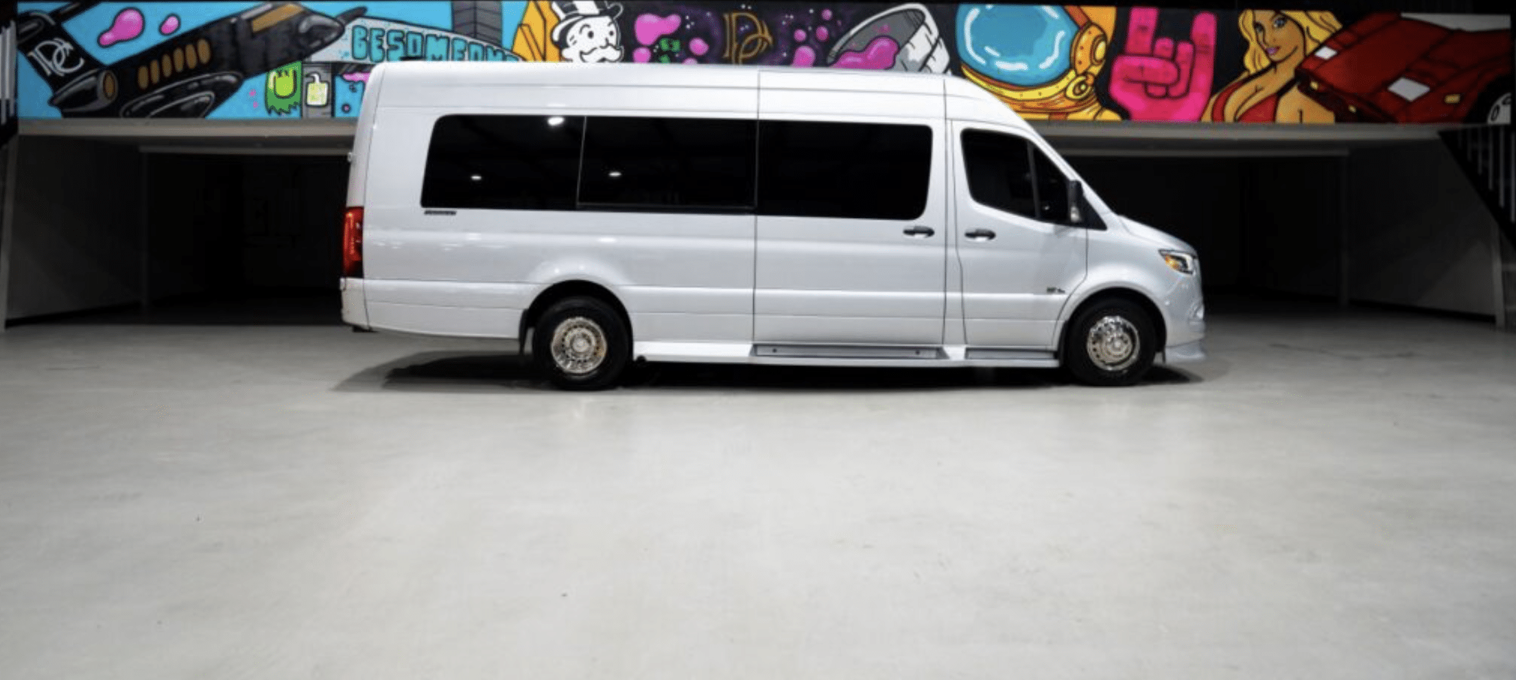 A white passenger van is parked indoors in front of a wall with colorful graffiti art, featuring cartoon-like characters and vibrant designs. The space appears clean and well-lit.